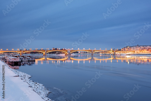 Winter twilight view of the illuminated Margaret Bridge with snow on the riverbank and reflection on the Danube River in Budapest, Hungary