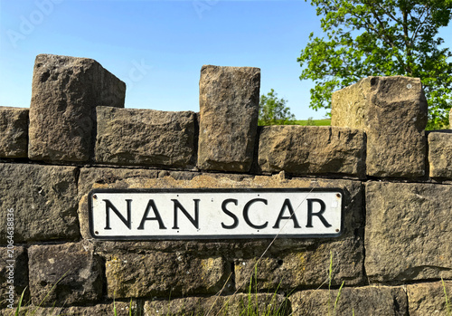 A traditional stone wall features a sign that reads 'Nan Scar'. Sunlight casts distinct shadows on the rough stone surface with greenery visible in the background in Cowling, Yorkshire, UK