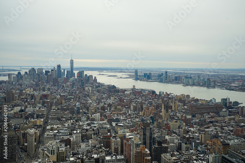 Manhattan cityscape sprawling towards the Hudson River, featuring the iconic One World Trade Center dominating the skyline, with the Statue of Liberty visible in the distance under an overcast sky