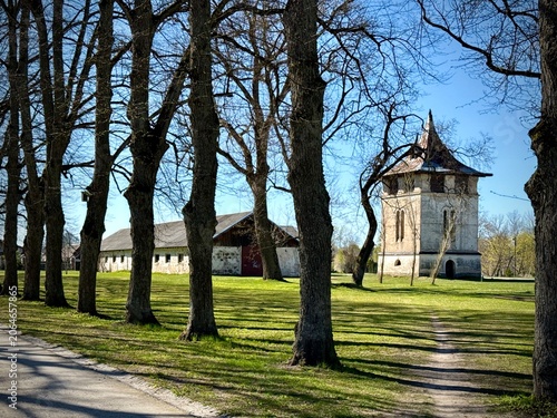 A historical water supply tower stands in a grassy field, flanked by tall, leafless trees. The tower features a rustic design with arched windows and a steep, pointed roof. 