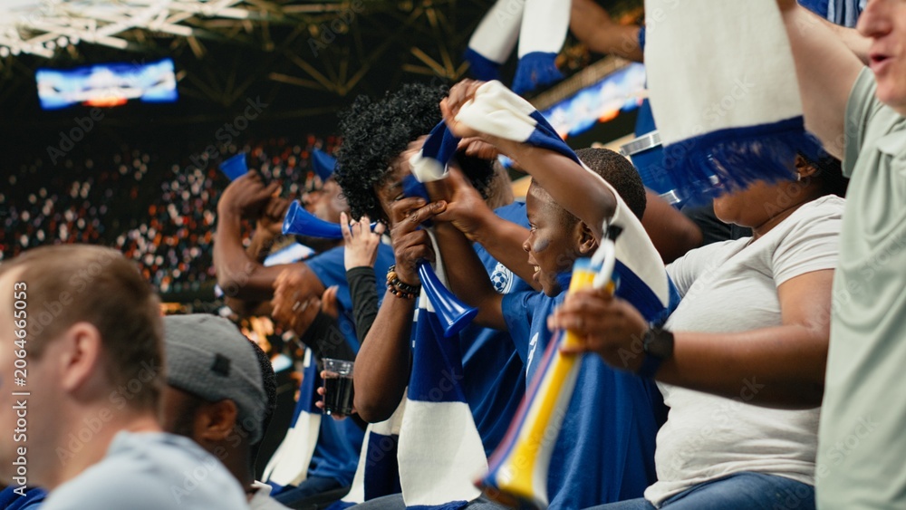 Obraz premium Black family celebrating with young child during exciting soccer match in packed stadium, cheering for blue team, parents sharing excitement of live sports with child fan, childhood joy.