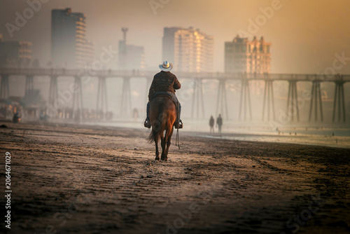 Behind view of a  cowboy riding a horse on a beach with a pier and buildings in the backgound