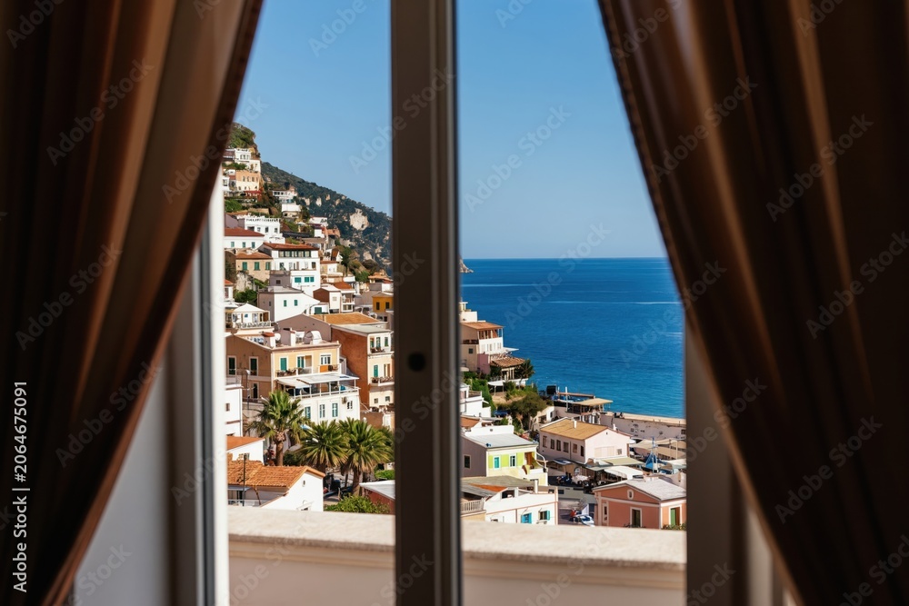 Naklejka premium View from open window to colorful buildings on seaside hill and blue ocean. Daytime sunny weather offers bright coastal landscape. Curtains frame scenic Mediterranean town.