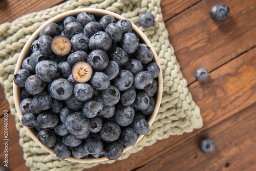 A high angle close up view of a round ceramic bowl filled with fresh ripe blueberries on a textured green placemat over a rustic dark brown wood surface