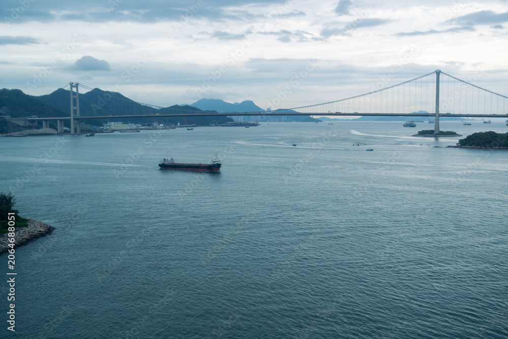 Fototapeta premium Large cargo ship sailing under suspension bridge on calm water during cloudy day with distant mountains