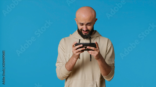 Canvas-taulu Joyful muslim man playing mobile video games in studio, enjoying online competition using smartphone app, with arabic gamer having fun with roleplaying challenge contest