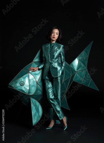 Fashion portrait of an Asian woman in a metallic teal pantsuit. Elegant model leaning on a large geometric origami fish sculpture against a black background