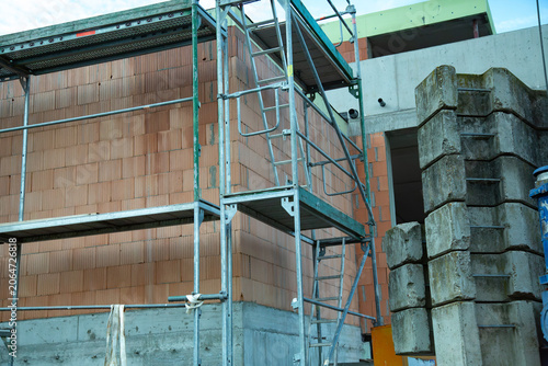 This professional outdoor photograph captures a modern construction site featuring temporary metal scaffolding installed next to a new building. The structure shows a newly built wall made of orange