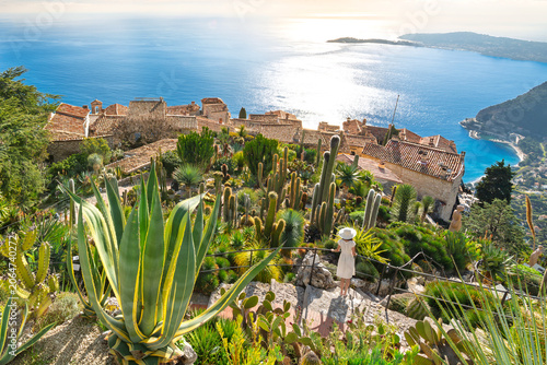 A female tourist stands on a narrow stone staircase of the botanical garden looking down on the medieval hilltop village of Eze Village, on the Mediterranean Coast of Southern France.