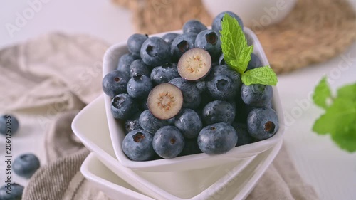 A crisp, high-angle close-up featuring a square white bowl overflowing with fresh, dew-covered blueberries and a vibrant mint garnish. Soft, natural lighting enhances the rich textures and cool tones 