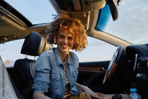 Schilderij op canvas Woman in casual denim jacket sits inside a car, smiling warmly, capturing a candid lifestyle moment with natural daylight and upbeat mood