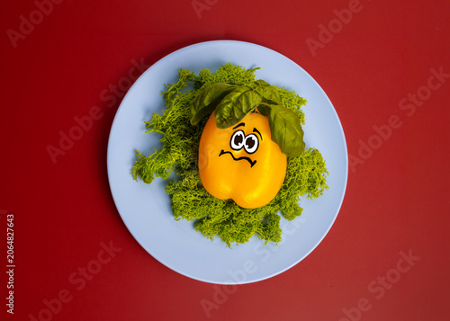 Yellow bell pepper character with sad painted face on green moss served on blue plate over red background flat lay