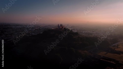 Aerial drone slowly approaches the Cholula pyramid and church at dawn, silhouetted against a magenta and blue sky with the Puebla city skyline and La Malinche volcano visible in the distance