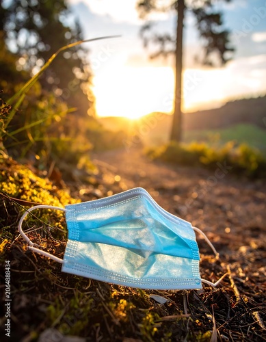 A discarded blue surgical mask lies on a dirt path surrounded by grass and trees at sunrise