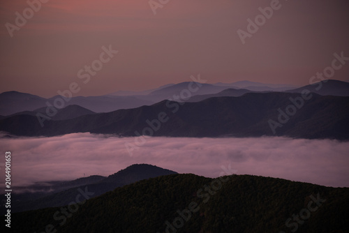 A breathtaking sunrise captured from Craggy Pinnacle near Asheville, North Carolina, with layers of rolling clouds filling the valleys below and warm light illuminating the Blue Ridge Mountains