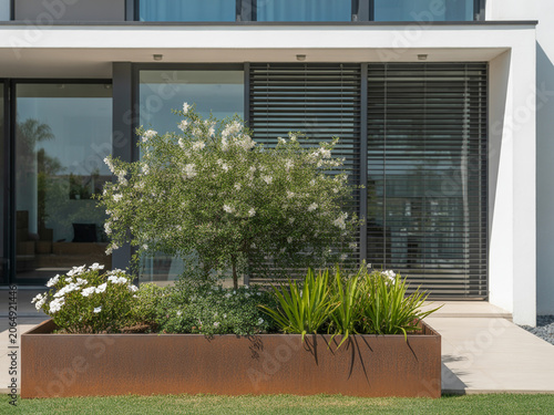 Modern Corten Steel Planter Box Featuring Lush Greenery and White Flowers Outside a Contemporary Residential Building