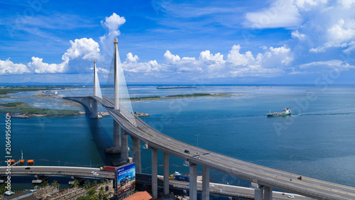 Cebu City, Philippines - 12 June 2026: Aerial view of the Cebu-Cordova Link Expressway bridge spanning the Mactan Channel with a cargo ship on the blue water under a cloudy sky.