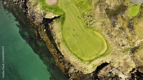 Aerial top-down view of people playing golf on a scenic green situated on a rugged, rocky coastline next to the turquoise ocean.