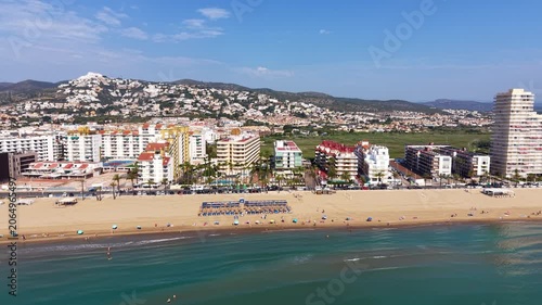 Peniscola beach coastline in Spain with turquoise Mediterranean sea, sandy shore, sunbathers, resort hotels, apartment buildings and green hills in background