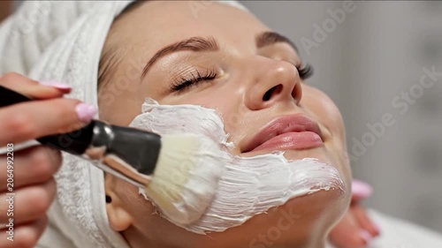Young woman receiving a white facial mask application with a cosmetic brush in a professional spa setting