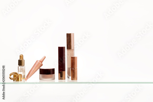 Collection of luxury cosmetic products including serum, lipstick, cream, and foundation bottles arranged on a glass shelf with minimal design for modern beauty and skincare concepts.