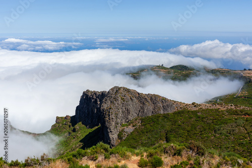 Mountain peak shrouded in misty clouds at Pico do Areeiro. Dramatic cliffs rise above lush highland terrain. Scenic hiking trail leads through Portugal's rugged landscape.
