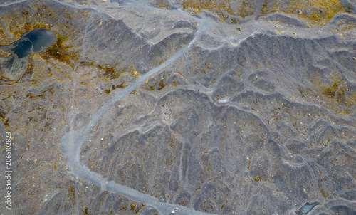 Drone aerial of Winding path crossing barren, rocky, and volcanic landscape. Small puddles of mossy water scatter across the textured ground