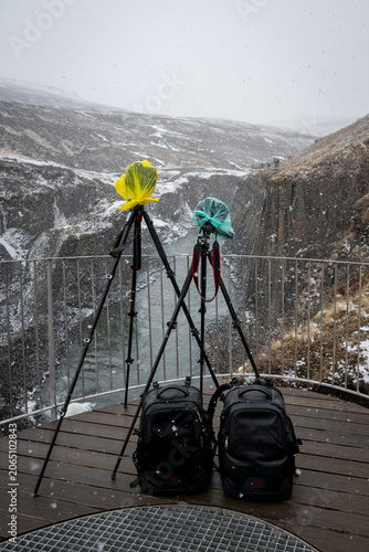 Tripods with rain covered cameras and backpacks on a viewing platform overlooking a snowy canyon with a river. Studlagil canyon Iceland