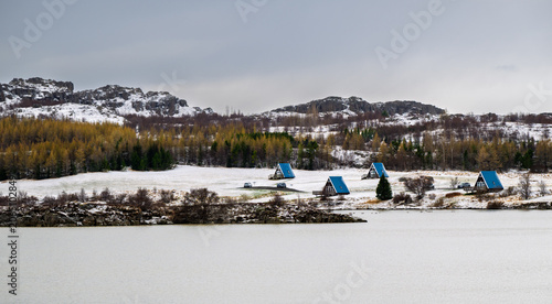 A frame holiday cabins with bright blue roofs sitting by a calm lake, surrounded by snow and distant mountains in the icelandic winter landscape. Iceland winter