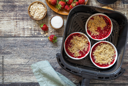 Homemade dessert traditional English crumble cake with strawberry in kitchen gadget modern air fryer on wooden background. Lifestyle trendy of new cooking. Top view flat lay background. Copy space.