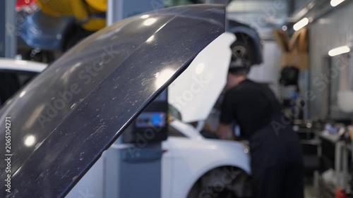 Close-up of the open hood of a car at a service station, with a blurry image of a mechanic in the background working on the car.