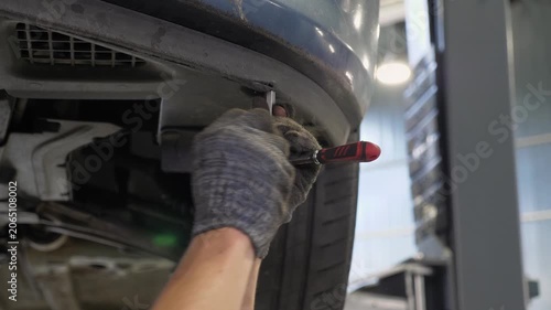 An auto mechanic is working under the car on a car lift in the garage. He disassembles the bottom of the car to repair it. Antifreeze is drained at the service station.