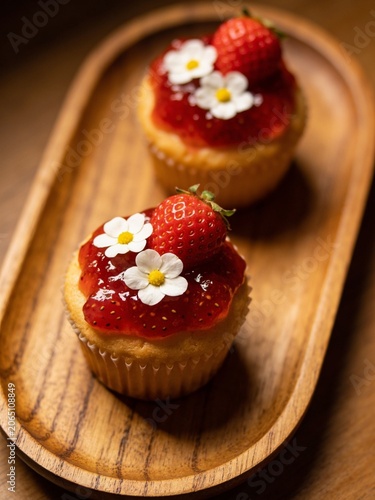 A beautifully composed commercial photograph featuring two homemade cupcakes topped with glossy red strawberry jam, fresh whole strawberries, and delicate white blossoms. Arranged elegantly on a rusti
