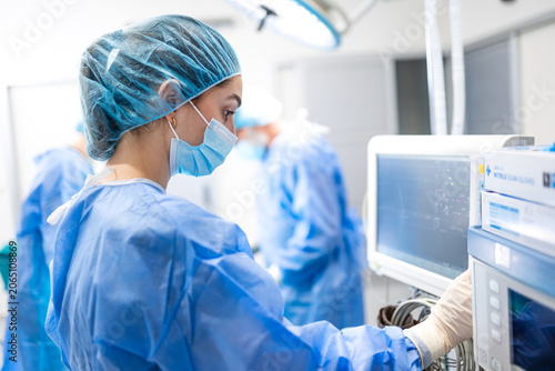 Female anesthesiologist wearing blue scrub cap and face mask adjusts anesthesia monitor in operating room, side profile view, surgical team blurred in background during hospital procedure preparation.