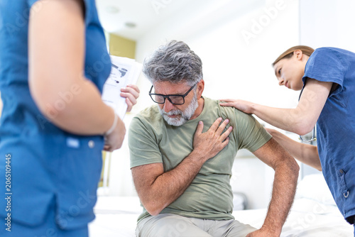 A concerned nurse and doctor provide care to an older male patient experiencing chest pain on a hospital bed. The team monitors the patient while reviewing medical documentation 