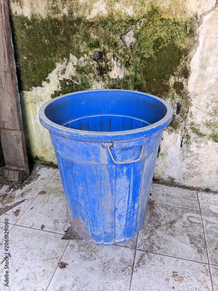 Fototapeta premium Large blue plastic bucket or trash bin placed outdoors against a mossy concrete wall.
