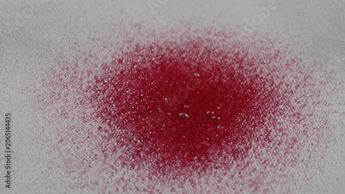 Viscous red liquid spreads across a white surface, transitioning from a droplet to a larger stain, showcasing the flow and texture of the substance in a close-up view