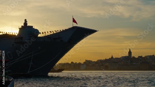 A massive grey naval vessel bow dominates the foreground in Istanbul, set against a golden sunset skyline and the historic cityscape of the Bosphorus strait