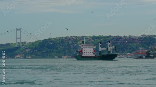 A large green cargo vessel navigates the Bosphorus strait in Istanbul, with a massive suspension bridge and lush green hills in the background under a clear sky