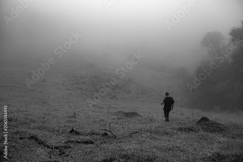 Obraz na plátně Atmospheric black and white landscape of lonely person walking into dense fog on grassy hillside evoking isolation and uncertainty for psychological advertising