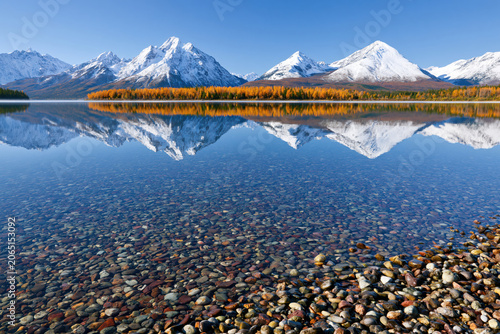 a serene landscape features a pristine lake with transparent water a bed of smooth, multi colored pebbles. above the waterline, the lake acts as a mirror, reflecting majestic snow covered mountains.