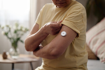 Senior woman adjusting wearable glucose sensor on upper arm at home, managing diabetes and remote health tracking. Useful for telehealth, chronic care, patient education marketing