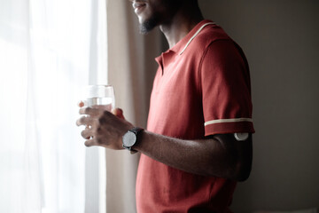 Young adult Black man holding glass of water near window, showing glucose sensor on arm and supporting diabetes self monitoring. Useful for healthcare campaigns, wellness apps, patient education