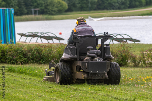 Groundskeeper Mowing Grass With Riding Lawn Mower Near Lake An unrecognizable groundskeeper mows grass with a riding lawn mower near a lake, showing park maintenance, landscaping service and summer ou