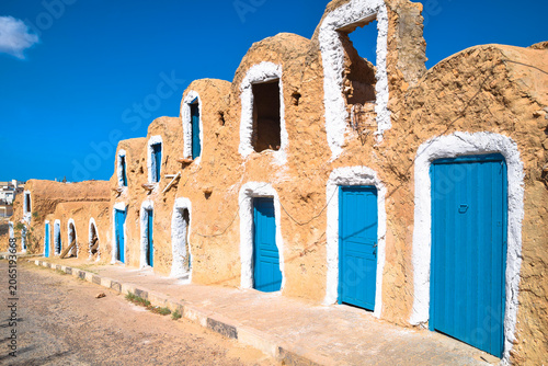 Traditional clay ghorfas with blue wooden doors and white windows along the street outside Ksar Metameur, Medenine, Tunisia.