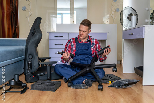 Photos Young man in uniform holding the gas lift piston cylinder and telescopic cover during the step by step assembly of a modern black gaming office chair