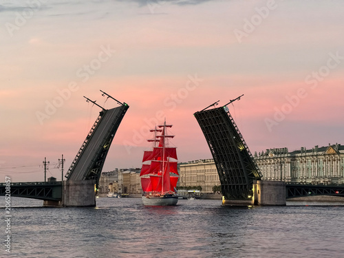 Fotografi Illuminated tall ship with beautiful red sails sailing under an open bascule bridge on a wide river during a vibrant sunset in a historic city