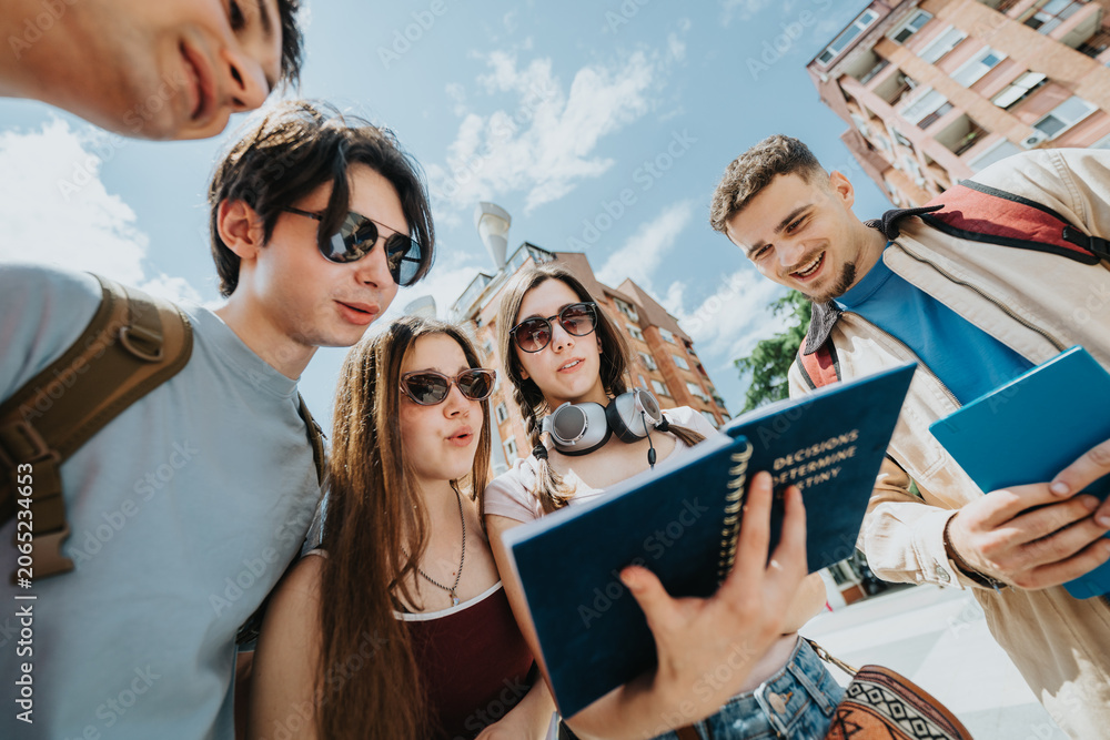 Fototapeta premium A group of students gather around a notebook while studying and discussing together outdoors. Young friends stand in front of campus buildings, reading notes and sharing ideas on a sunny day.