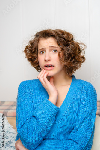 young woman with a shocked and worried expression touching her cheek, displaying concern and anxiety, with visible dental braces and curly hair indoors on a light background