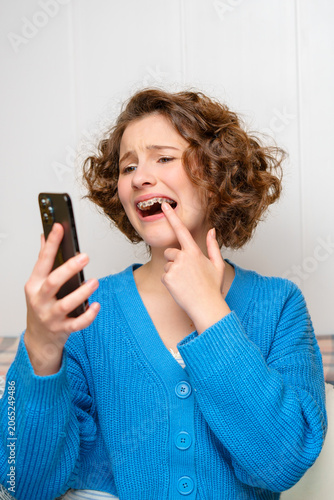 young woman with dental braces looking at her smartphone with a worried expression, pointing at her teeth, highlighting orthodontic discomfort and self-consciousness